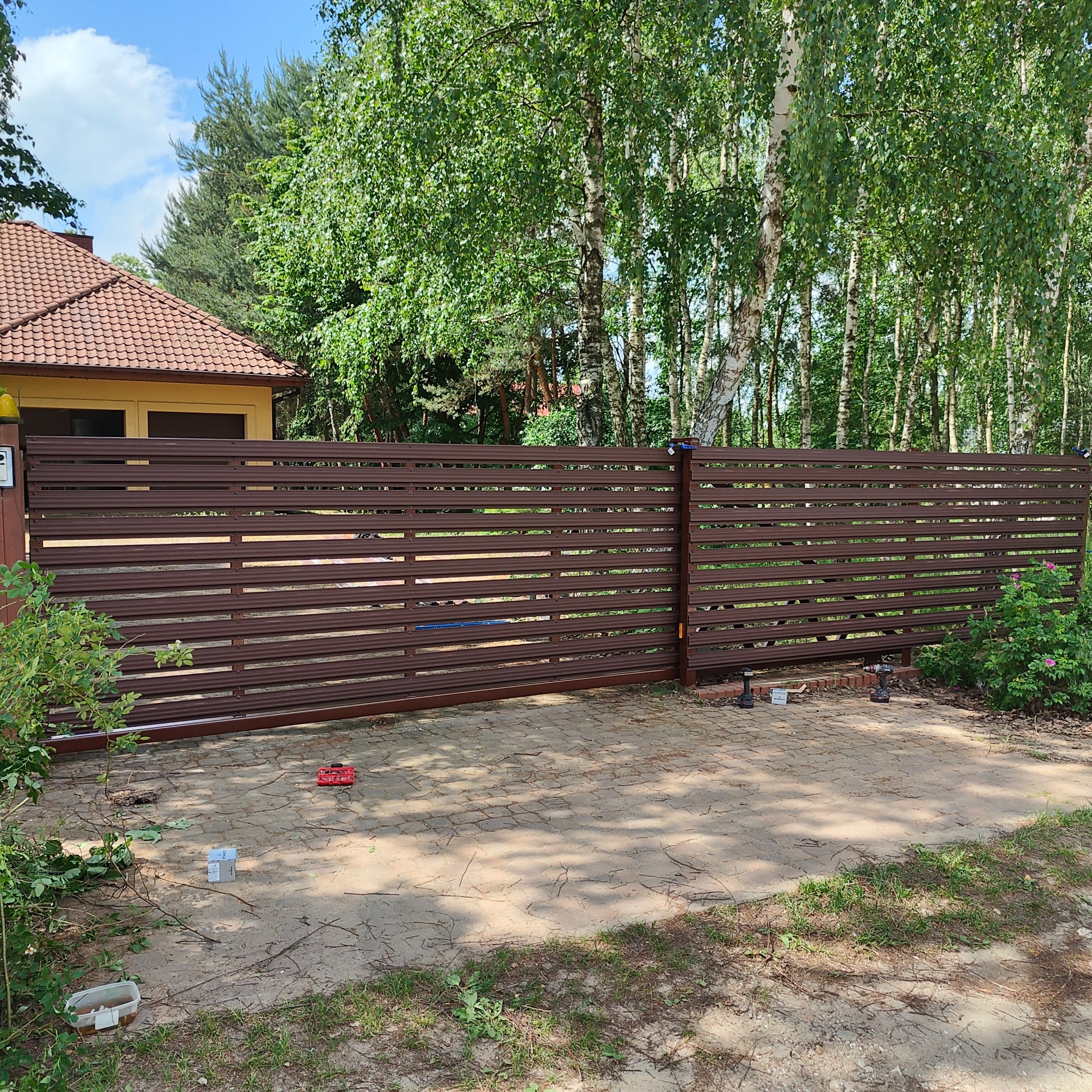 Nowoczesne, poziome ogrodzenie z brązowych paneli metalowych przed domem z czerwonym dachem, widoczne narzędzia montażowe i otaczająca zieleń.