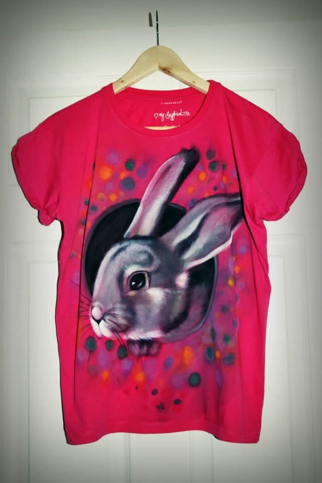Różowa koszulka z ręcznie malowanym portretem szarego królika, wisząca na wieszaku na tle białych drzwi. Widoczne kolorowe kropki wokół portretu.