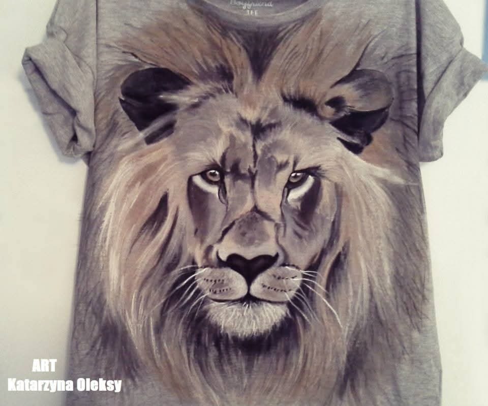 Szara koszulka z ręcznie malowanym portretem lwa, widoczne detale sierści i wyraziste oczy. Sygnowana w lewym dolnym rogu.