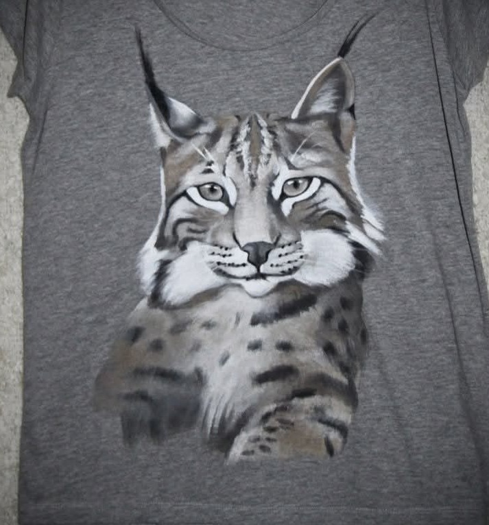 Szara koszulka z ręcznie malowanym portretem rysia w odcieniach szarości i bieli, leżąca na płaskiej powierzchni. Widoczne detale sierści i charakterystyczne uszy rysia.