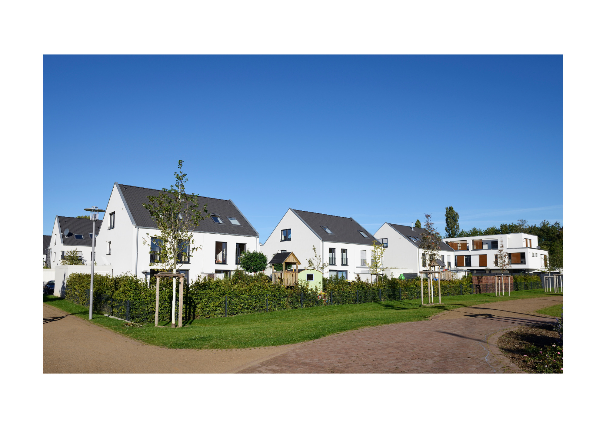 Osiedle nowoczesnych, białych domów jednorodzinnych z ciemnymi dachami, otoczone zielenią i młodymi drzewkami, widok z poziomu ulicy.