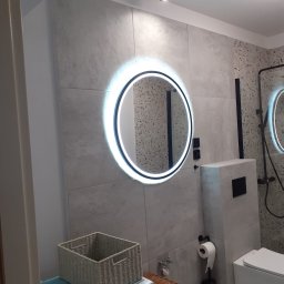 Adamix Adam Grzebielucha - Nowoczesna łazienka z okrągłym lustrem LED, szarą armaturą i płytkami imitującymi kamień, kosz z wikliny na pralce.