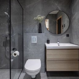 Adamix Adam Grzebielucha - Nowoczesna łazienka z szarymi marmurkowymi płytkami, kabiną prysznicową typu walk-in z czarną ramą, wiszącą toaletą, umywalką z drewnianą szafką i okrągłym lustrem.