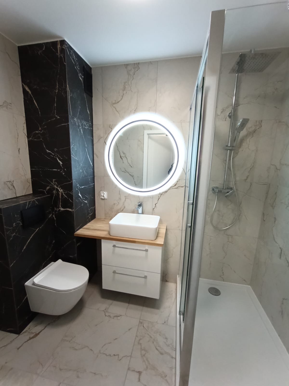 Nowoczesna łazienka z marmurowymi płytkami, okrągłym lustrem z podświetleniem LED, wiszącą toaletą i kabiną prysznicową z deszczownicą.