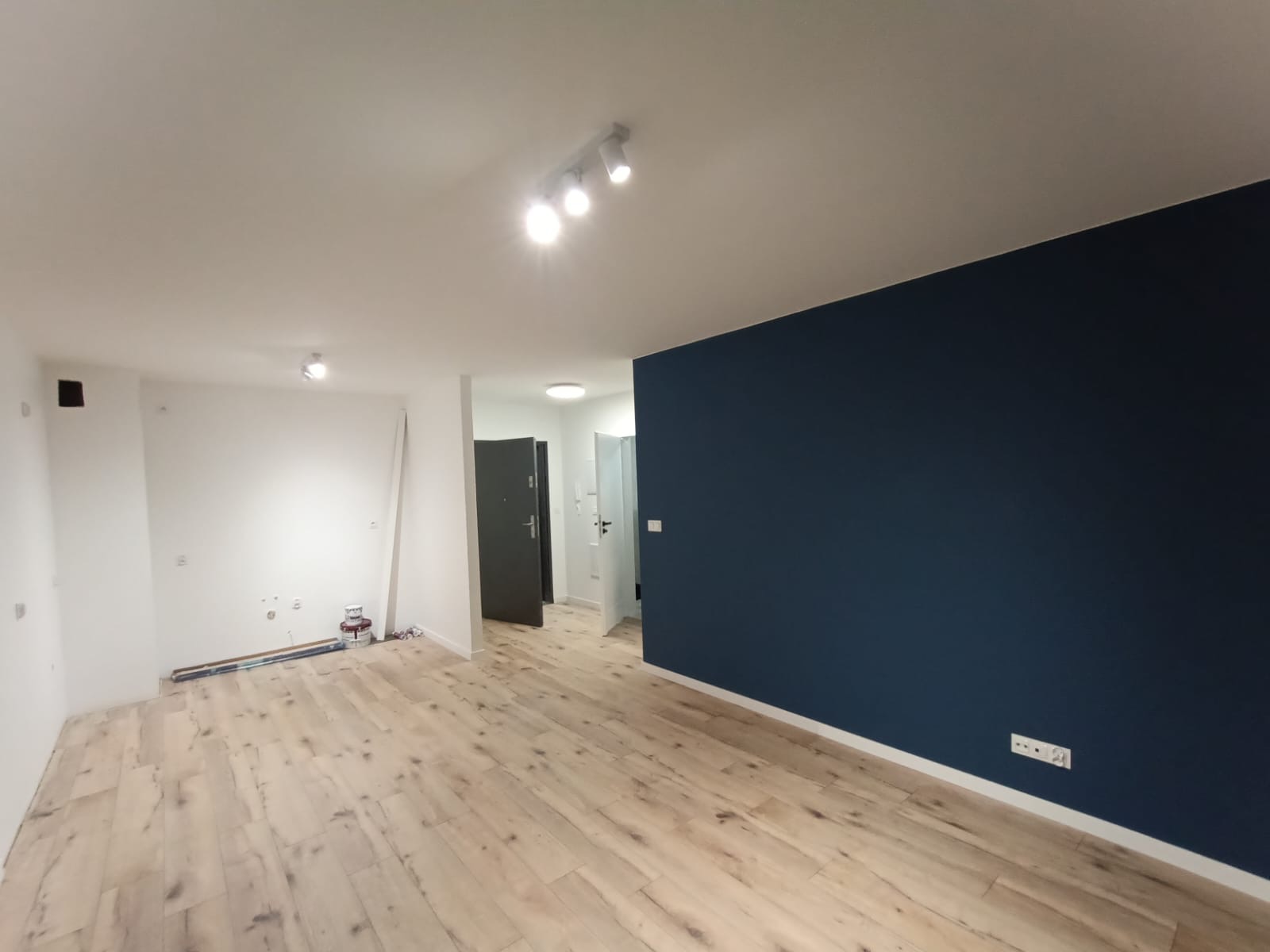 Wnętrze po remoncie: parkiet w odcieniach jasnego drewna, jedna ściana pomalowana na granatowo, pozostałe białe, widoczne puszki z farbą przy ścianie, oświetlenie sufitowe punktowe.