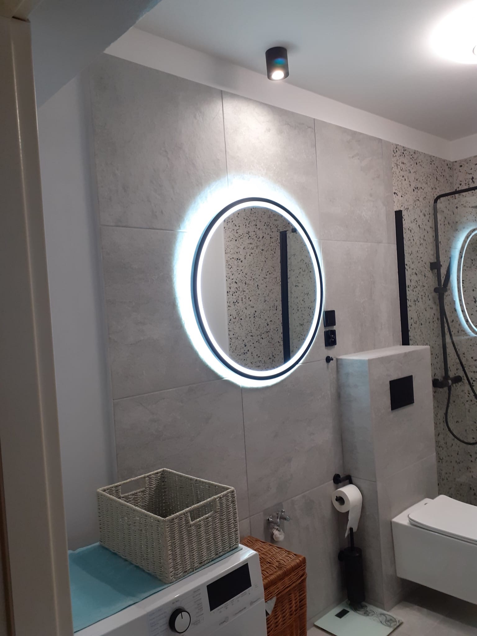 Nowoczesna łazienka z okrągłym lustrem LED, szarą armaturą i płytkami imitującymi kamień, kosz z wikliny na pralce.