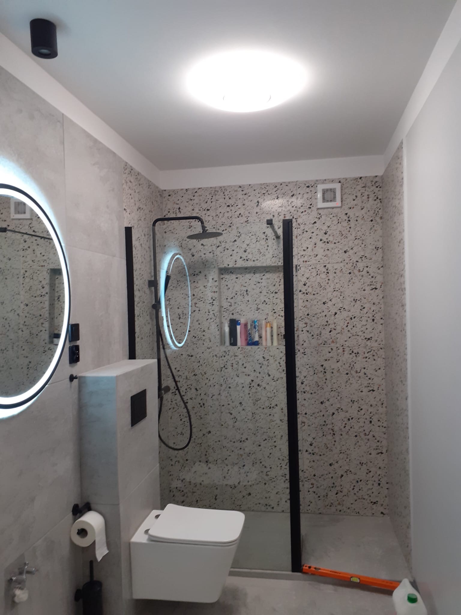 Nowoczesna łazienka z kabiną prysznicową typu walk-in, okrągłym lustrem z podświetleniem LED, podwieszaną toaletą i ścianami wyłożonymi płytkami imitującymi lastryko; na podłodze leży poziomica...