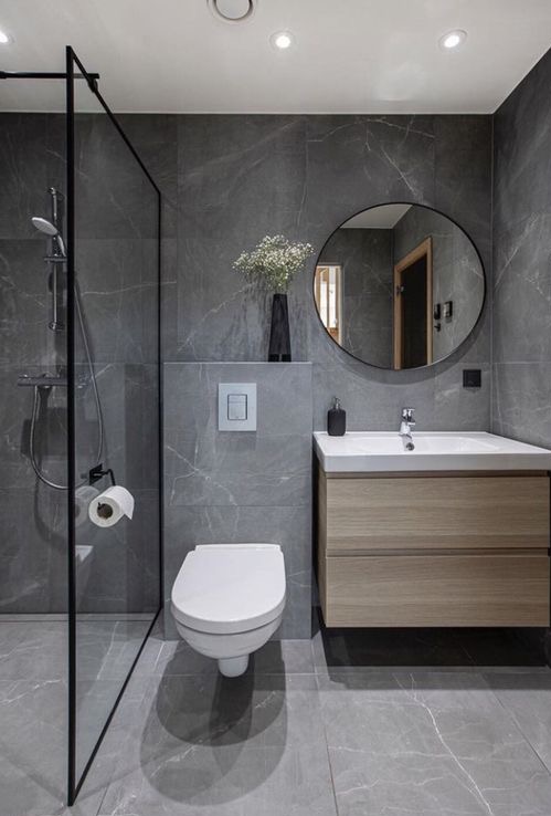 Nowoczesna łazienka z szarymi marmurkowymi płytkami, kabiną prysznicową typu walk-in z czarną ramą, wiszącą toaletą, umywalką z drewnianą szafką i okrągłym lustrem.