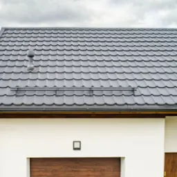 Nowoczesny dom z dachem pokrytym szarą dachówką metalową, widoczny system rynnowy i komin wentylacyjny. Na pierwszym planie fragment białej elewacji z drewnianą bramą garażową i drzwiami wejściowymi.