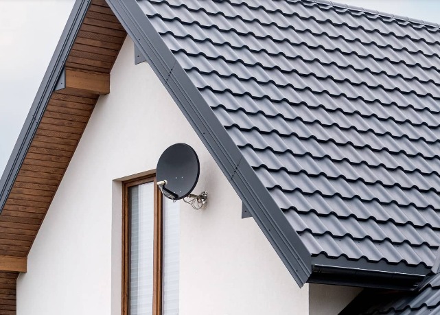 Fragment domu z nowym, grafitowym dachem z dachówki stalowej, widoczna antena satelitarna na elewacji, okno z brązowymi ramami i drewniane podbitki dachowe.