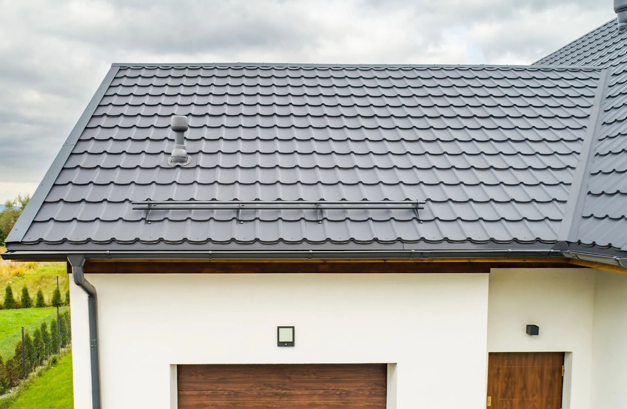 Nowoczesny dom z dachem pokrytym szarą dachówką metalową, widoczny system rynnowy i komin wentylacyjny. Na pierwszym planie fragment białej elewacji z drewnianą bramą garażową i drzwiami wejściowymi.