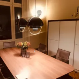 Jadalnia z drewnianym stołem, krzesłami i nowoczesnymi lampami wiszącymi z okrągłymi elementami świetlnymi, na ścianie biała szafa z dekoracyjnymi świecznikami, widok z niskiego kąta.