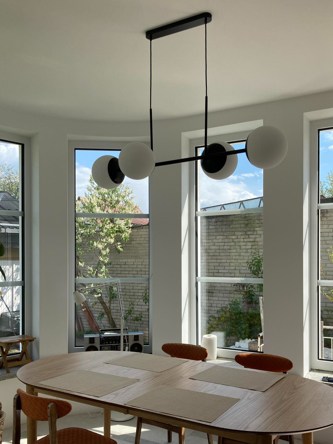 Nowoczesna lampa wisząca z trzema kulistymi kloszami nad drewnianym stołem w jasnej jadalni z dużymi oknami wychodzącymi na ogród.