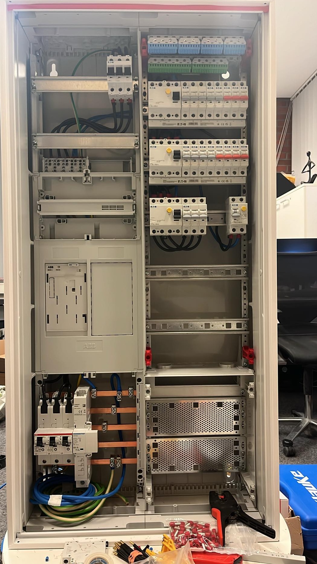 Rozdzielnica elektryczna w trakcie montażu, widoczne przewody, zabezpieczenia, listwy zaciskowe, oraz narzędzia i końcówki kablowe na dole szafy.