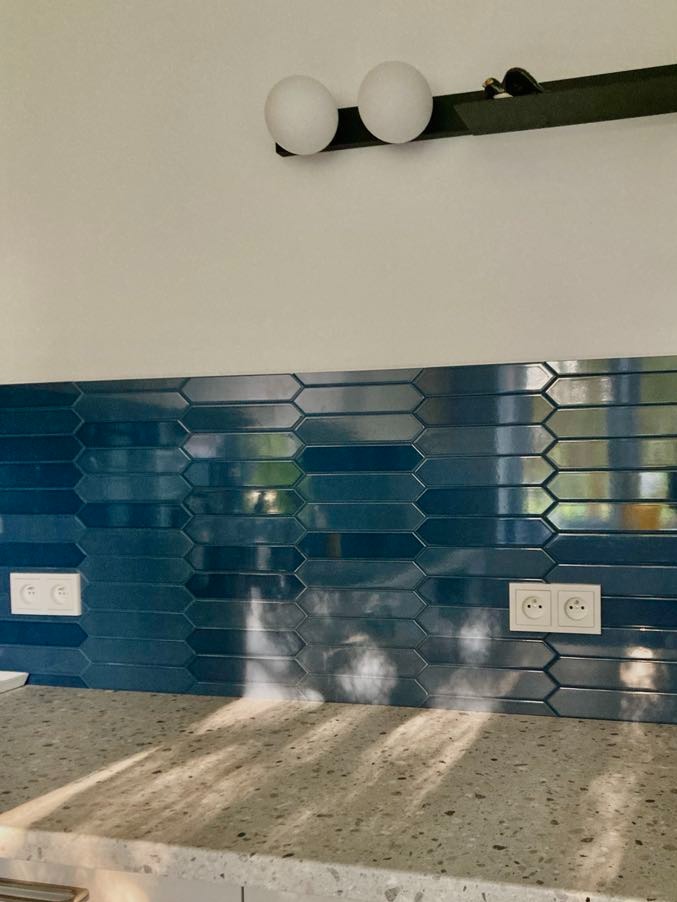 Błyszcząca, niebieska mozaika ułożona na ścianie nad blatem, z widocznymi dwoma podwójnymi gniazdkami elektrycznymi i lampą z dwoma kulistymi kloszami na czarnej listwie.