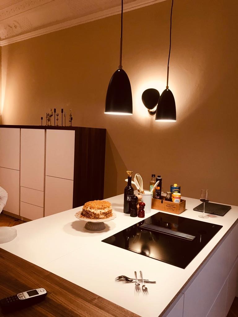 Nowoczesna kuchnia z białym blatem, czarną płytą indukcyjną z pochłaniaczem oparów i wiszącymi lampami w kształcie dzwonków. Na blacie ciasto na paterze, przyprawy i butelka wina. Na pierwszym...