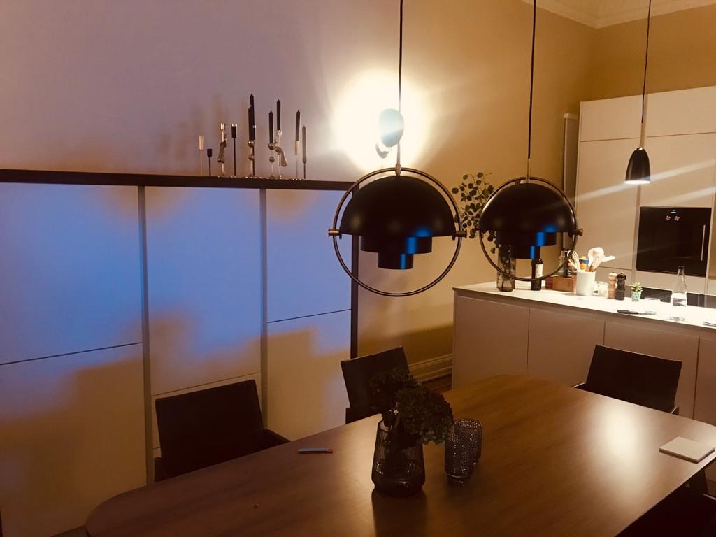 Jadalnia z drewnianym stołem, czarnymi krzesłami i trzema nowoczesnymi lampami wiszącymi z czarnymi kloszami, biała zabudowa kuchenna w tle, dekoracyjne świeczniki na szafce.