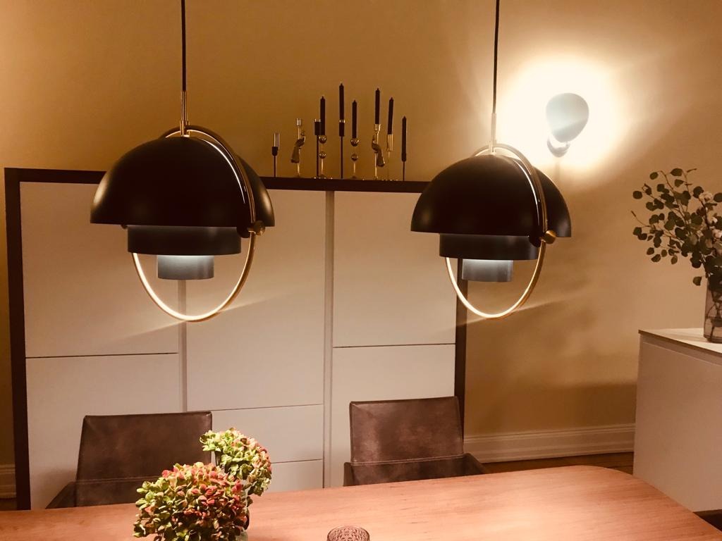 Dwa czarne, designerskie wiszące plafony z elementami w kolorze złotym oświetlają stół w jadalni, nad nim dekoracyjne świeczniki na komodzie i rośliny w wazonie.