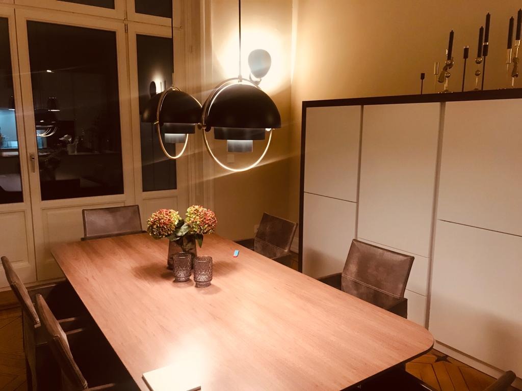 Jadalnia z drewnianym stołem, krzesłami i nowoczesnymi lampami wiszącymi z okrągłymi elementami świetlnymi, na ścianie biała szafa z dekoracyjnymi świecznikami, widok z niskiego kąta.