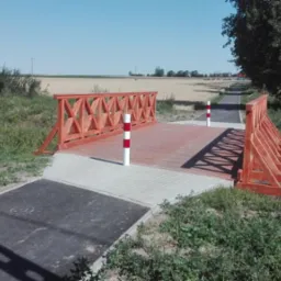 Drewniany mostek nad ścieżką rowerową, z czerwonymi barierkami o ażurowej konstrukcji i dwoma słupkami ostrzegawczymi, otoczony zielenią i polami w tle.