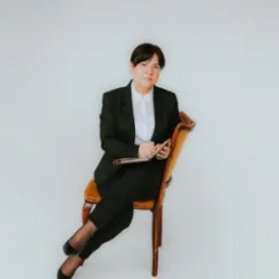 Kobieta w czarnym garniturze siedzi na krześle z notatnikiem i długopisem w rękach, spoglądając w obiektyw na jasnym tle.