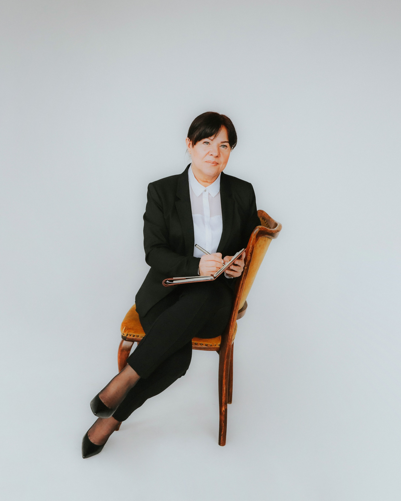 Kobieta w czarnym garniturze siedzi na krześle z notatnikiem i długopisem w rękach, spoglądając w obiektyw na jasnym tle.