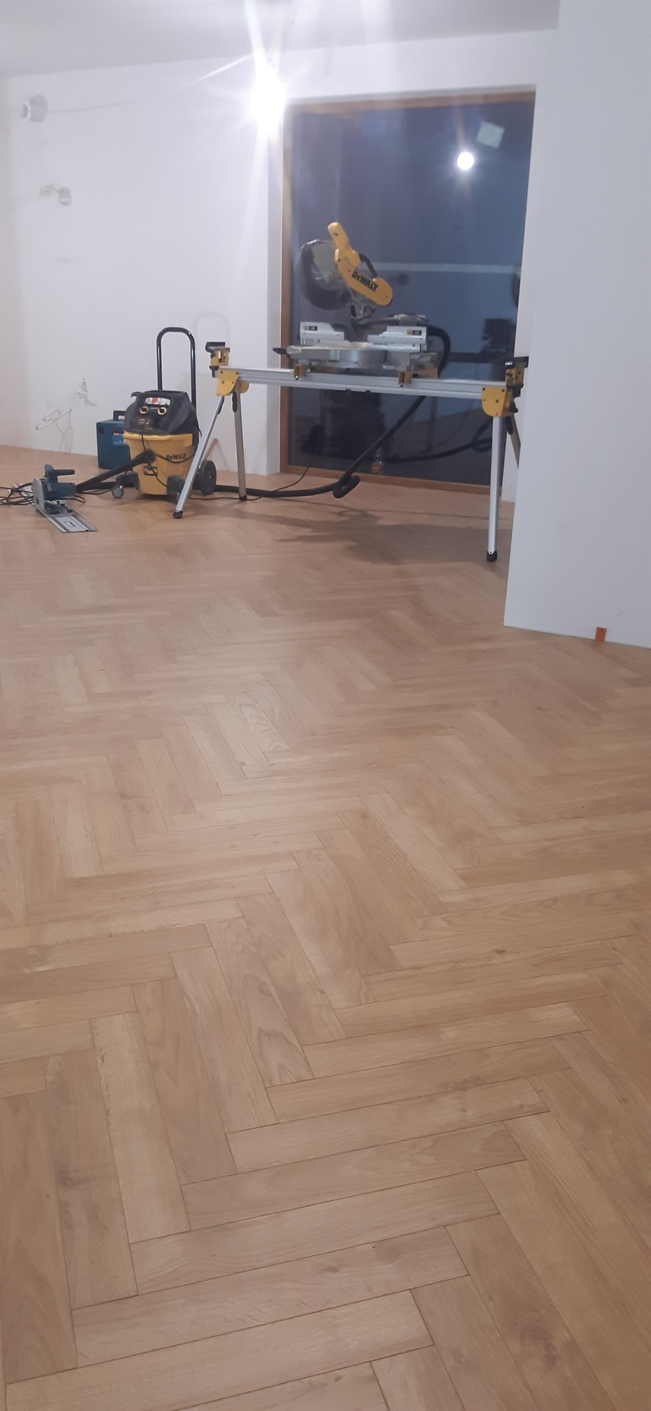 Jasna podłoga ułożona w jodełkę, widoczna piła stołowa DeWalt na stojaku, odkurzacz przemysłowy i listwa przypodłogowa w tle, w trakcie montażu.