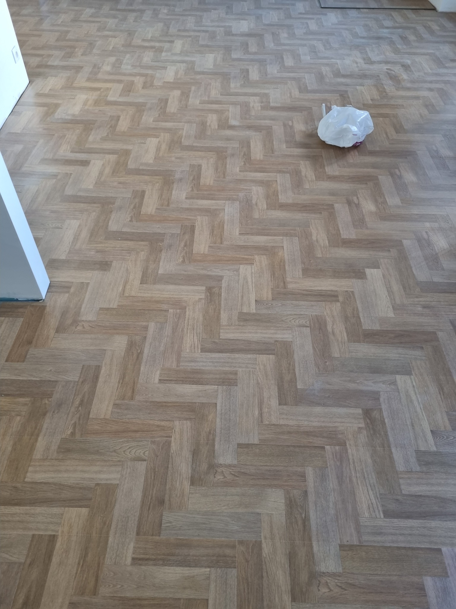 Ułożona podłoga z paneli w wzór jodełki, z widoczną białą plastikową torbą po prawej stronie i fragmentem białej ściany z lewej strony.