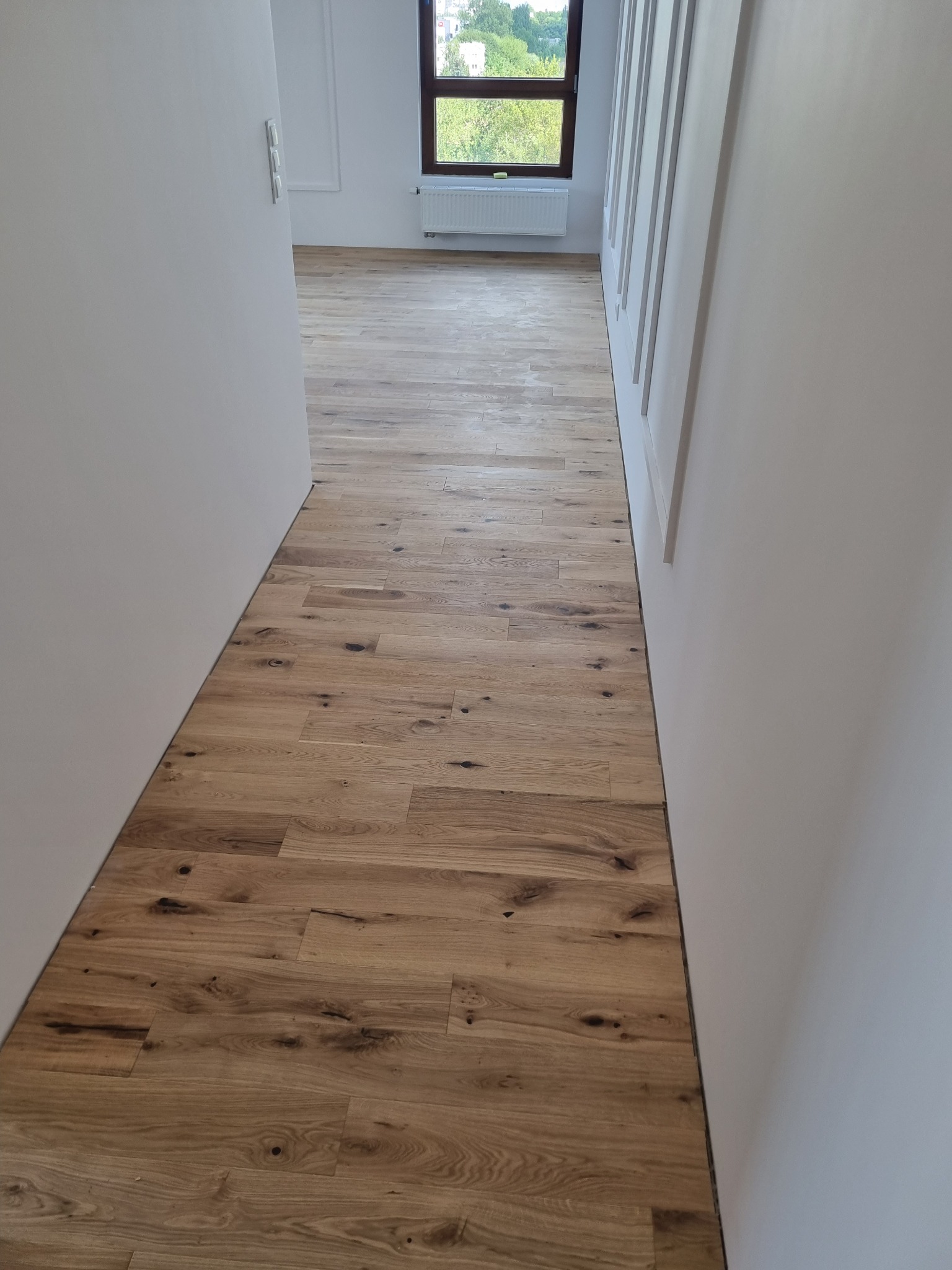 Korytarz z nową, jasną podłogą z desek drewnianych ułożonych wzdłuż, widok w kierunku okna z zielenią za szybą, białe ściany z dekoracyjnymi listwami.