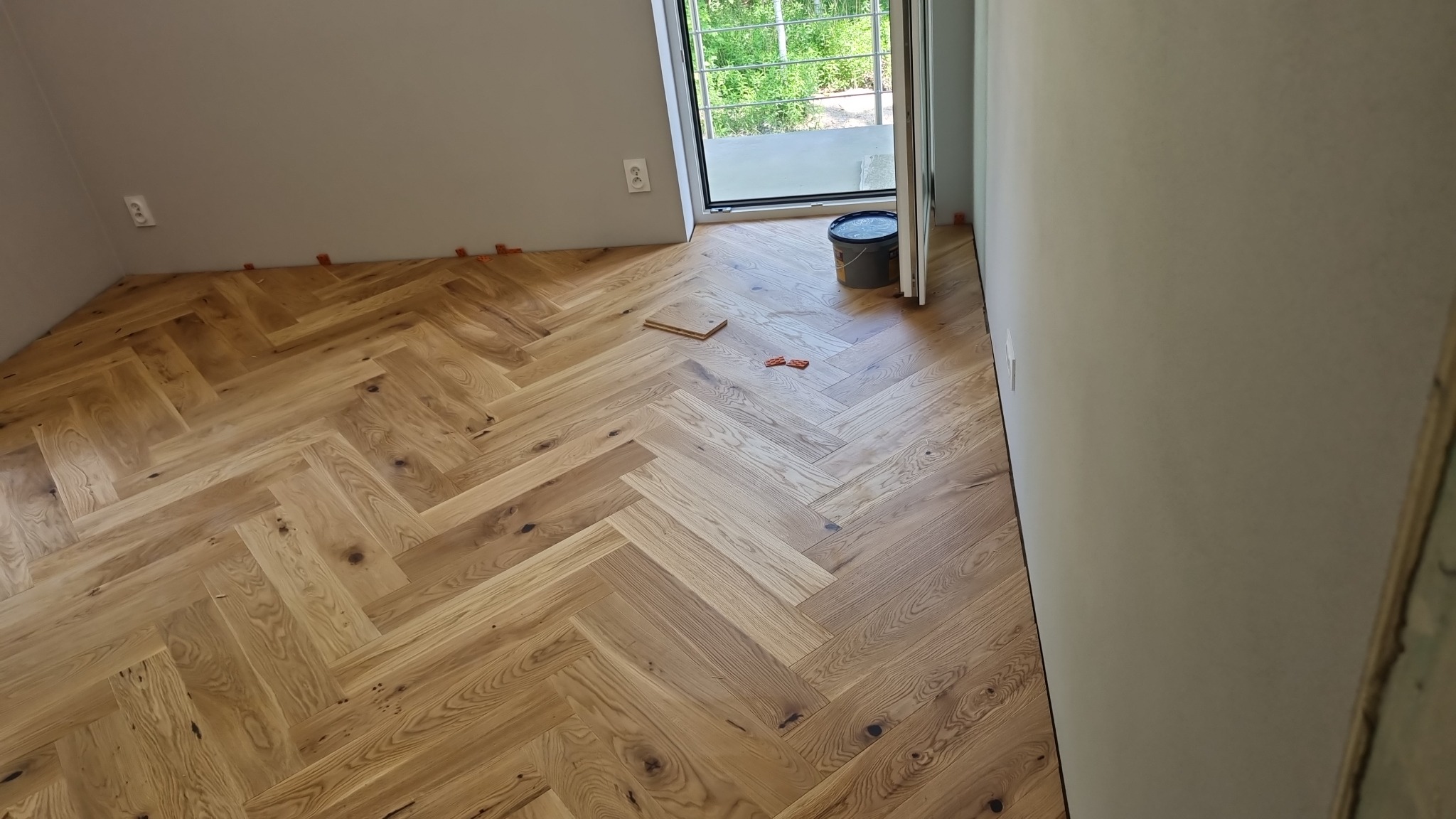 Jasny parkiet ułożony we wzór jodełki francuskiej w pokoju z oknem, widoczne podkładki dystansowe przy ścianie, wiaderko i kilka desek na podłodze.