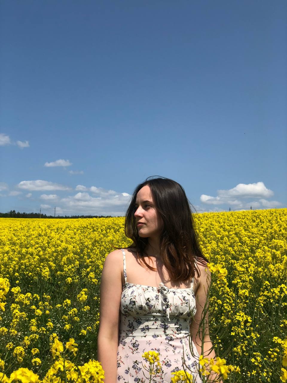 Kobieta w kwiecistej sukience stoi na skraju pola kwitnącego rzepaku, z błękitnym niebem i kilkoma chmurami w tle.