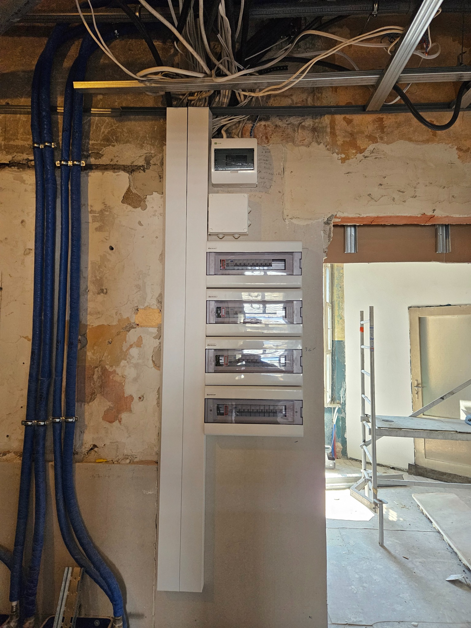 Rozdzielnica elektryczna z zabezpieczeniami w trakcie montażu na ścianie z widocznymi przewodami i elementami konstrukcyjnymi sufitu podwieszanego.