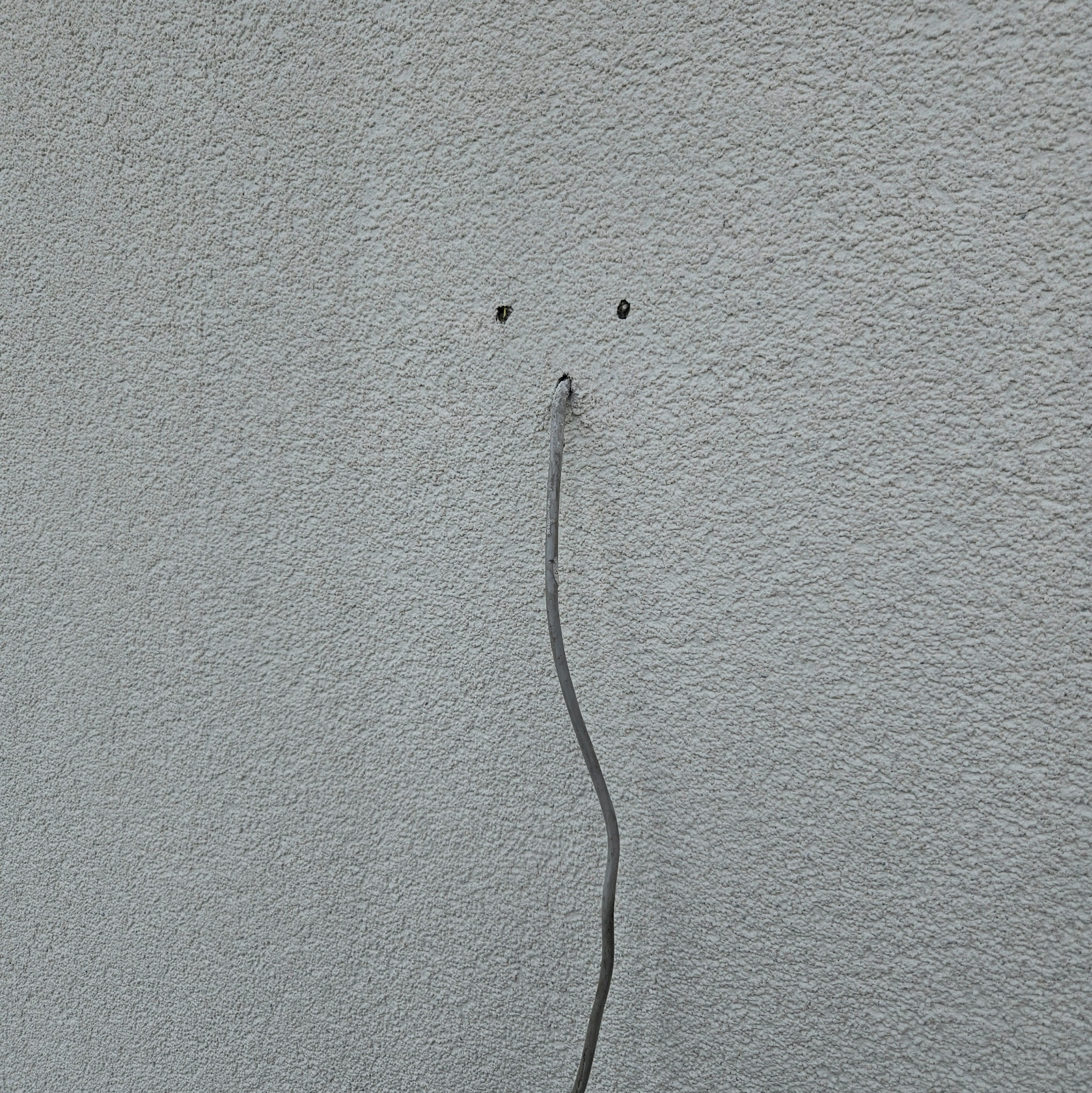 Szary kabel wystaje ze ściany o szorstkiej fakturze. Widoczne dwa otwory powyżej, sugerujące planowany montaż. Przewód elektryczny w trakcie instalacji.