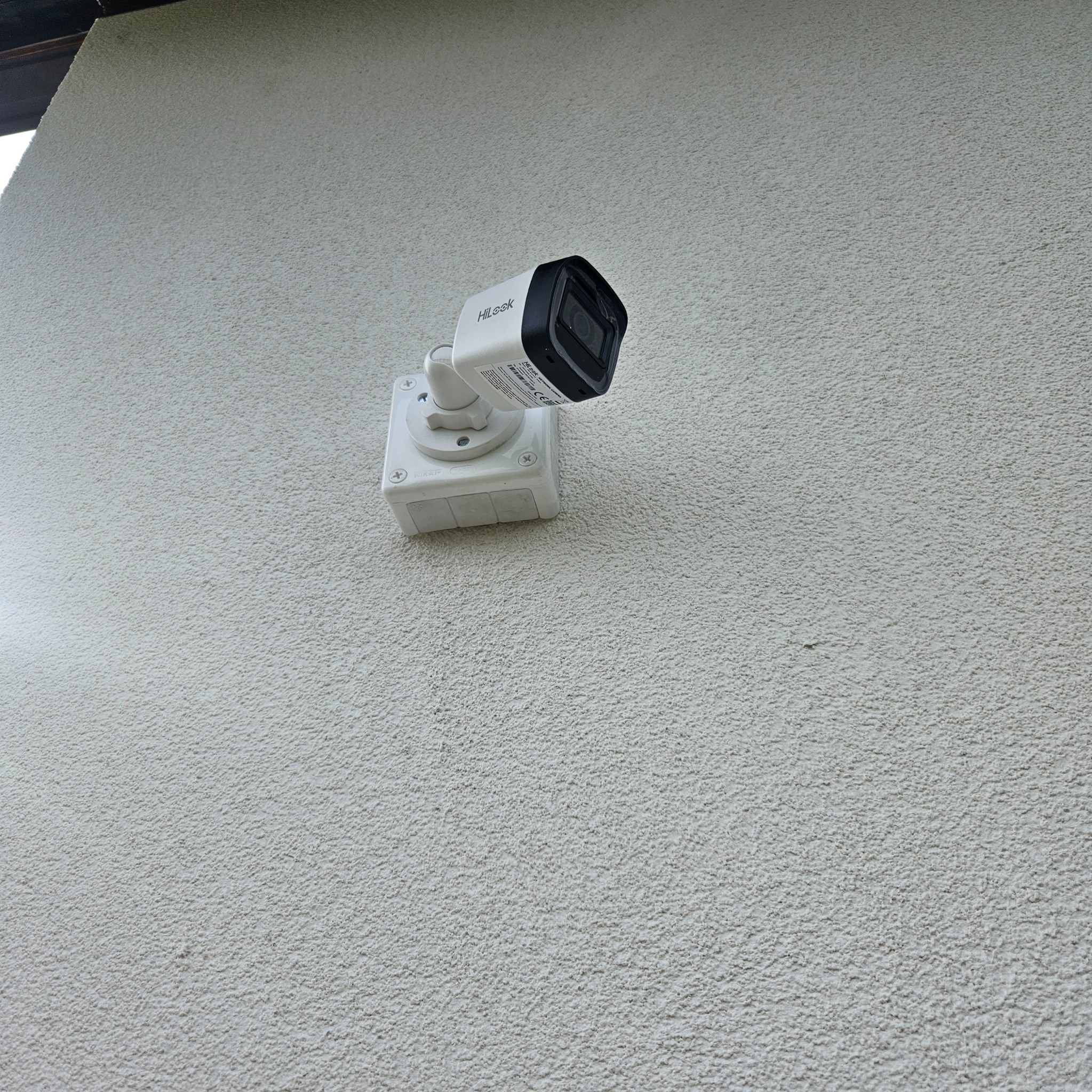 Biała kamera HiLook zamontowana na chropowatej, jasnej ścianie zewnętrznej. Widoczne śruby mocujące i fragment dachu w górnym, lewym rogu.