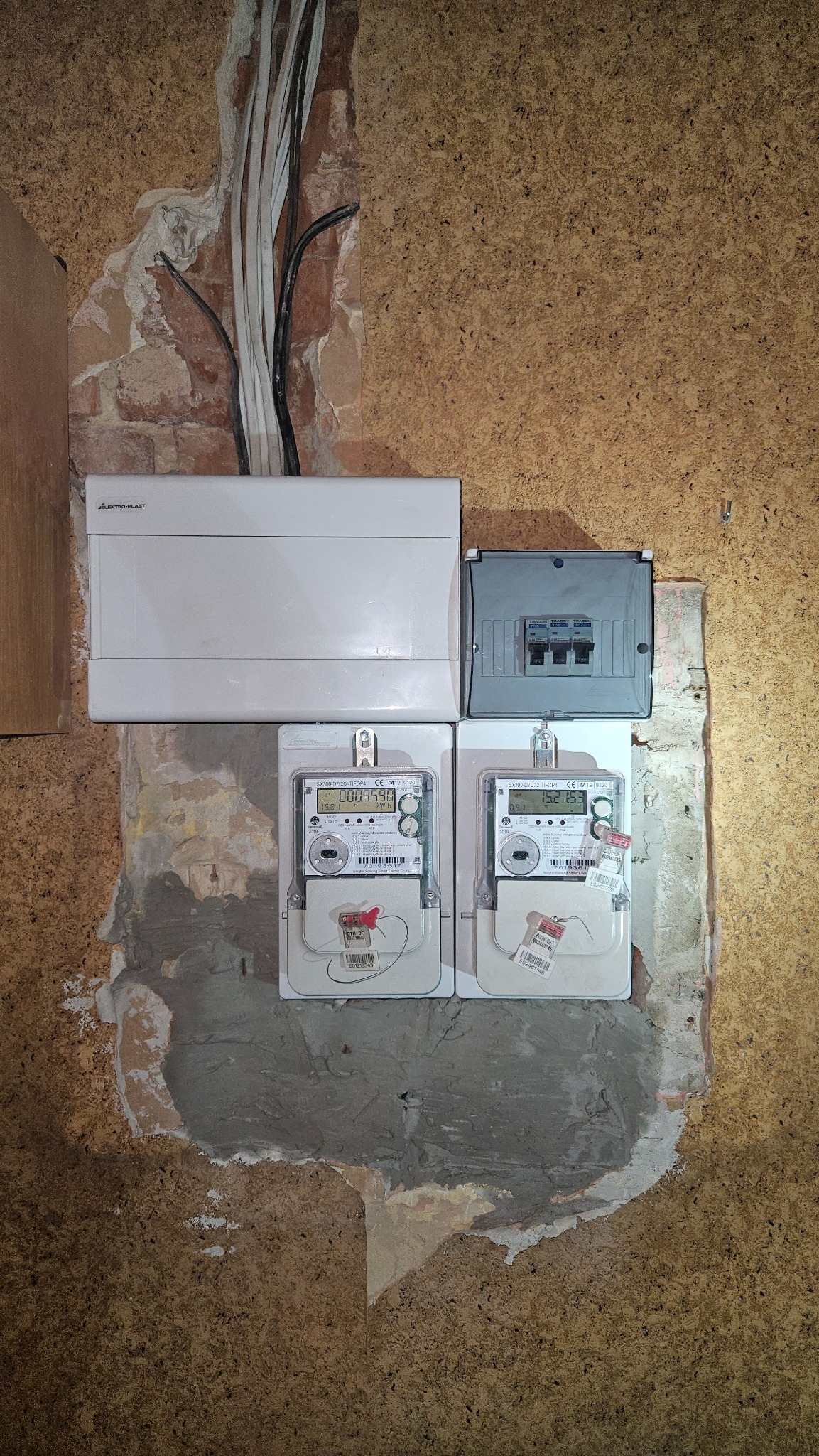 Dwa liczniki elektryczne zamontowane obok siebie na ścianie z widocznymi przewodami i skrzynkami rozdzielczymi. Widoczne ślady po remoncie.