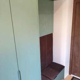 Pracownia Meblowa Modena - Szafa wnękowa z siedziskiem w odcieniach zieleni i brązu, z ryflowanym panelem dekoracyjnym. Siedzisko tapicerowane, widoczny fragment drewnianych drzwi i wycieraczki.