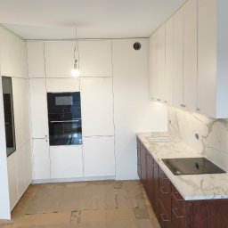 Pracownia Meblowa Modena - Biała zabudowa kuchenna z drewnianymi szafkami dolnymi i marmurowym blatem w Bratkowicach. Widoczny piekarnik i płyta indukcyjna. Podłoga zabezpieczona kartonem.