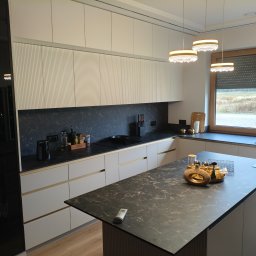 Pracownia Meblowa Modena - Nowoczesna kuchnia z wyspą, fronty szafek w odcieniach bieli i szarości, blat imitujący marmur. Nad wyspą dekoracyjne lampy. Widok na zewnątrz przez okno.