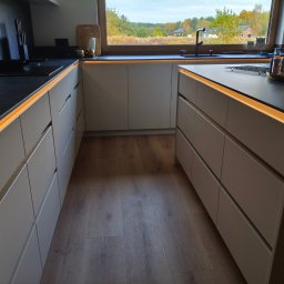 Pracownia Meblowa Modena - Nowoczesna kuchnia z szarymi szafkami i czarnym blatem, oświetlona listwami LED. Widok na okno z zielenią w tle. Minimalistyczny design, drewniana podłoga.