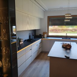 Pracownia Meblowa Modena - Nowoczesna kuchnia z szarymi szafkami, czarnym blatem i oświetleniem LED. Na wyspie kuchennej dekoracyjny zestaw. Za oknem widok na zewnątrz.