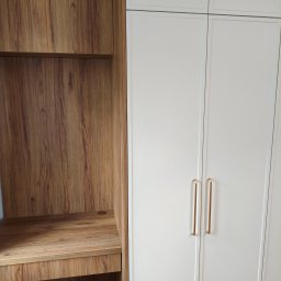 Pracownia Meblowa Modena - Biała szafa z frezowanymi frontami i złotymi uchwytami sąsiaduje z drewnianym regałem z biurkiem. Widoczny kabel zasilający w dolnej części regału.