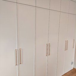 Pracownia Meblowa Modena - Biała szafa wnękowa z frezowanymi frontami i złotymi uchwytami, sięgająca sufitu, z widoczną kratką wentylacyjną w dolnej części i drewnianą podłogą.