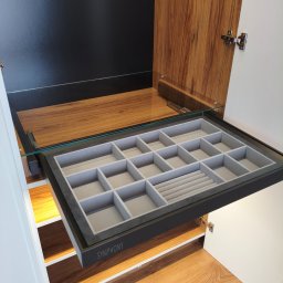 Pracownia Meblowa Modena - Wysuwana szuflada z przegródkami na biżuterię w szafie wnękowej. Szuflada z czarną ramą i szarym wnętrzem, widoczne drewniane elementy szafy i podłoga.