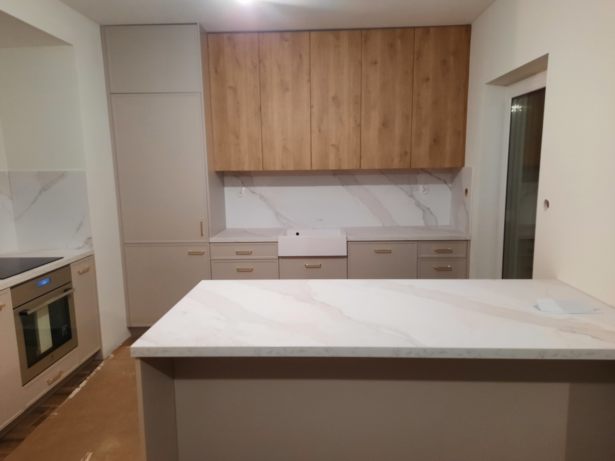 Nowoczesna kuchnia w Bratkowicach z beżowymi szafkami dolnymi i drewnopodobnymi górnymi, marmurowy blat wyspy kuchennej z widocznymi żyłami, piekarnik w zabudowie. Podłoga częściowo zabezpieczona...