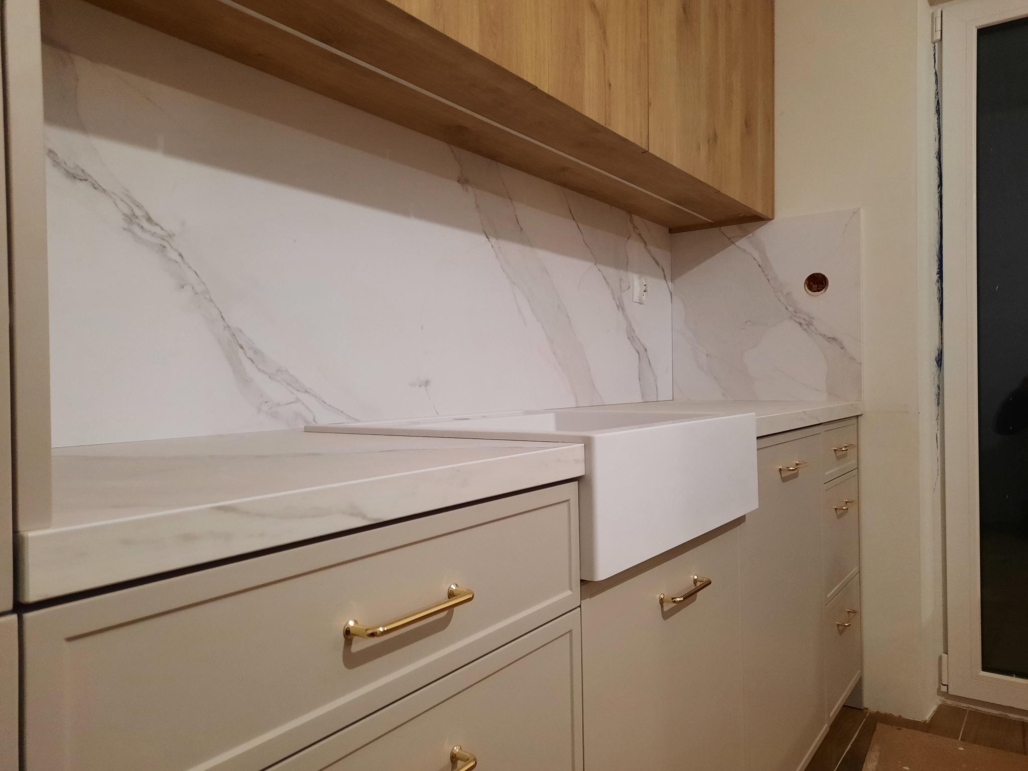 Beżowe szafki kuchenne z marmurowym blatem i białym zlewem farmerskim, złote uchwyty, marmurowa ściana nad blatem, drewniana szafka wisząca.