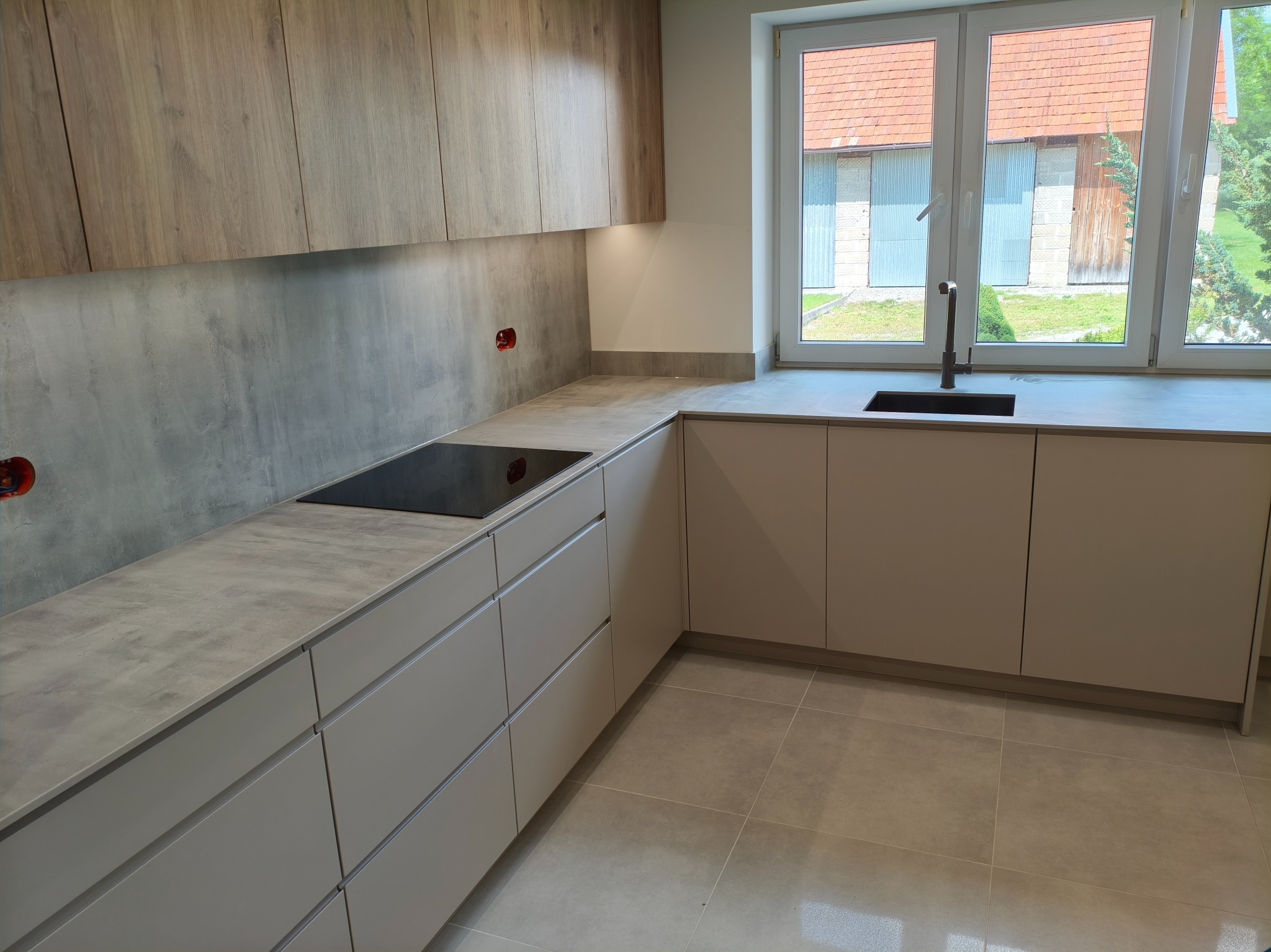 Nowoczesna kuchnia z drewnianymi szafkami górnymi i szarymi dolnymi, z blatem w kolorze betonu. Widok na okno z widokiem na budynek gospodarczy.