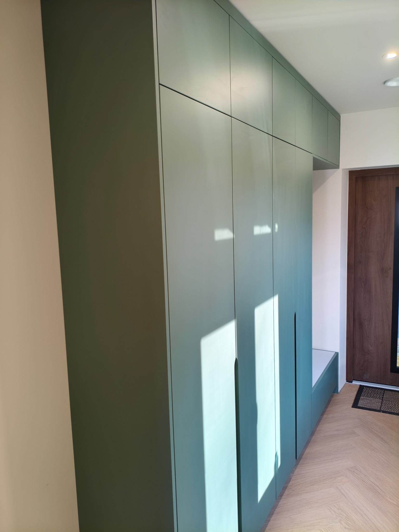 Długa, oliwkowa szafa wnękowa z siedziskiem, w przedpokoju z parkietem ułożonym w jodełkę. Minimalistyczny design, brak uchwytów, drzwi sięgają sufitu.
