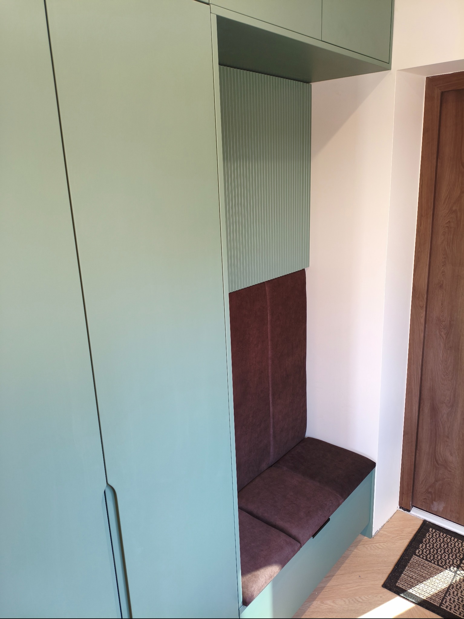 Szafa wnękowa z siedziskiem w odcieniach zieleni i brązu, z ryflowanym panelem dekoracyjnym. Siedzisko tapicerowane, widoczny fragment drewnianych drzwi i wycieraczki.
