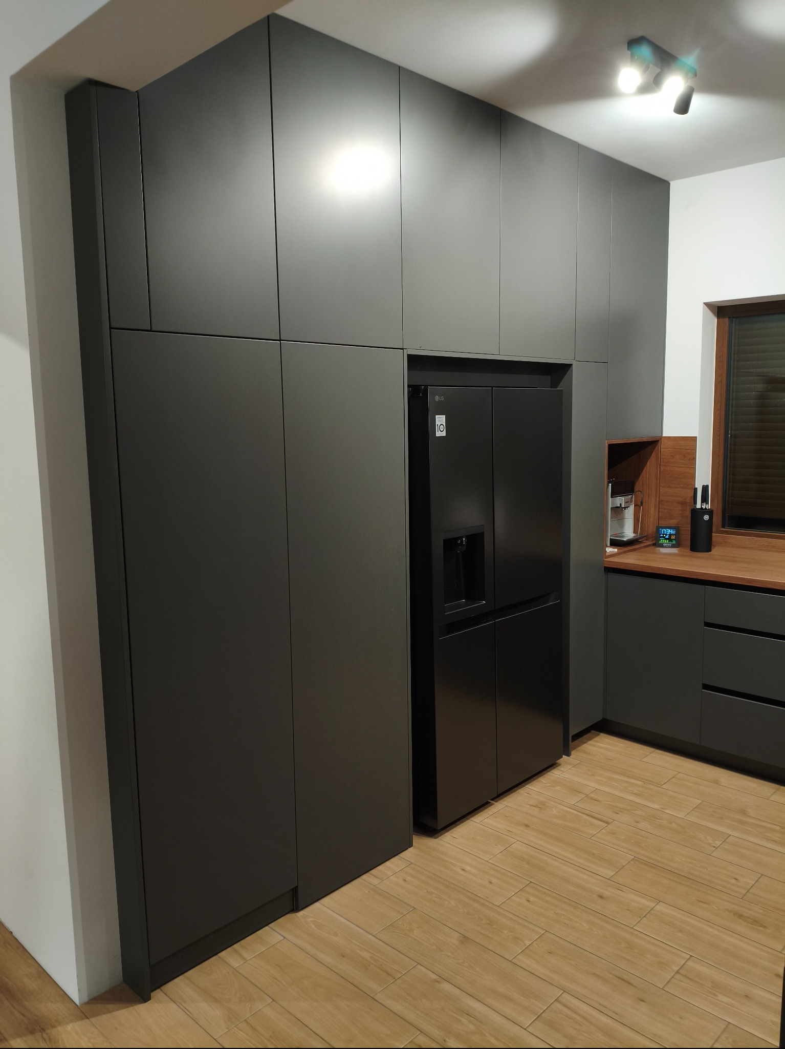 Nowoczesna kuchnia z zabudową w kolorze grafitowym, obejmującą lodówkę side-by-side. Jasna drewniana podłoga i blat dodają ciepła. Minimalistyczne oświetlenie sufitowe.