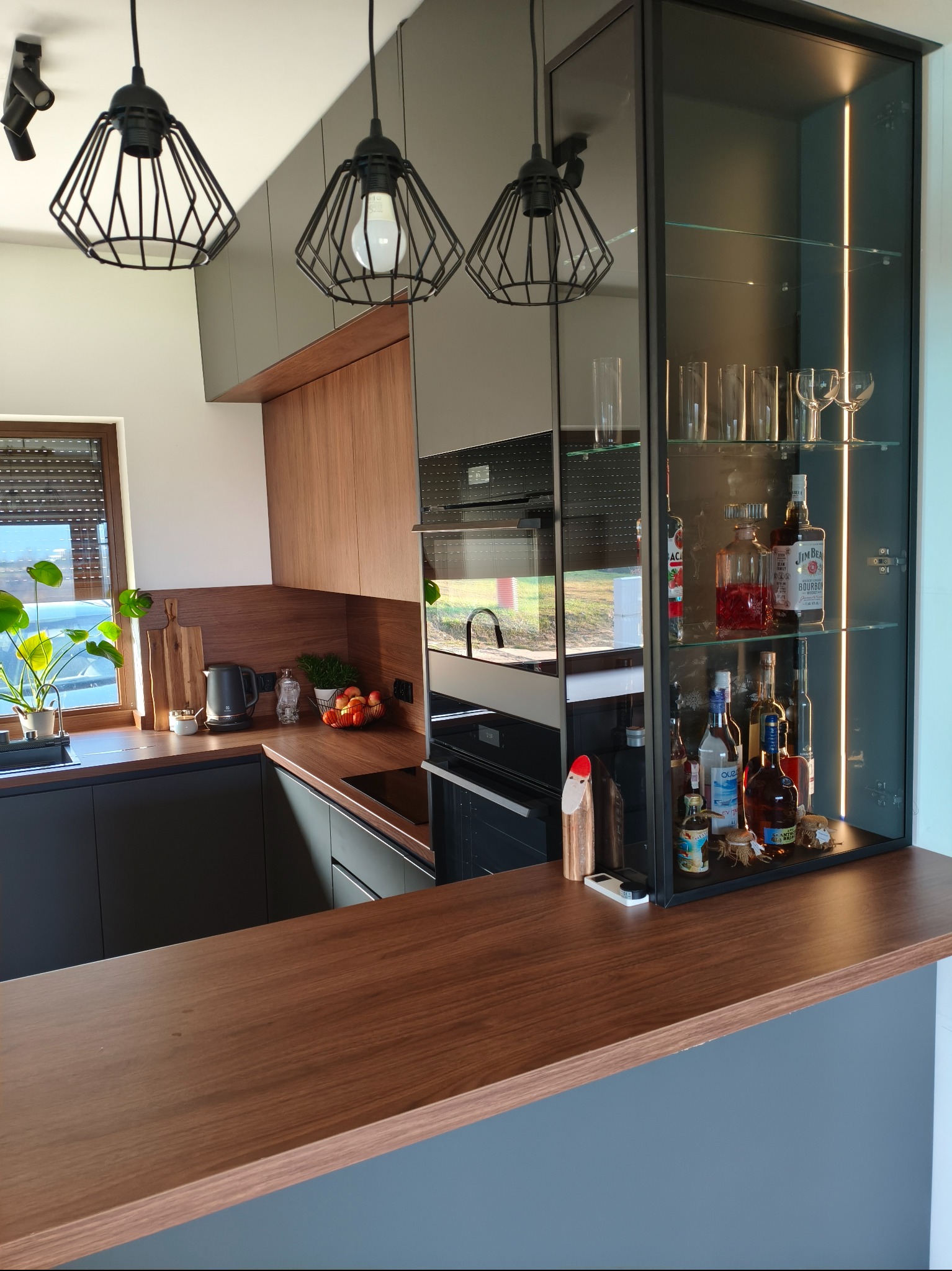 Nowoczesna kuchnia z drewnianymi frontami, czarnymi szafkami i przeszkloną witryną z oświetleniem LED, eksponującą alkohole i szkło. Uwagę przykuwają designerskie lampy.